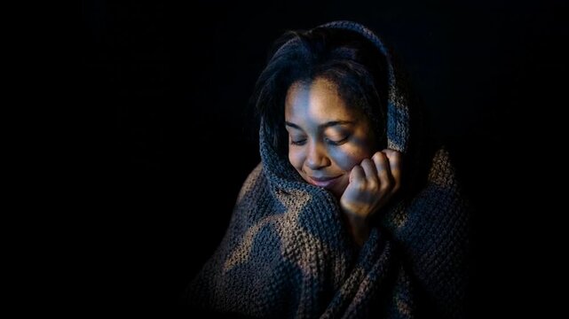 Young person wrapped in a textured blanket, gazing thoughtfully with light illuminating their face against a dark background