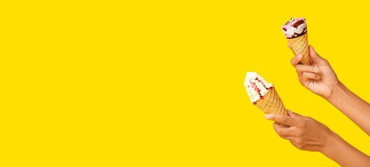 Fototapeta premium Two hands holding melting ice cream cones with strawberry and chocolate swirls on bright yellow background, the sweet dessert treats are presented against a bright yellow background