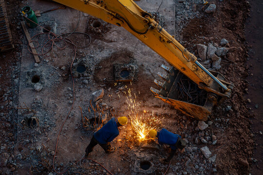 Top view of construction worker welding steel reinforcement structure for bridge foundation with excavator equipment during golden hour light