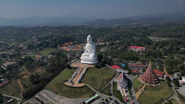 Aerial view of the Wat Huay Pla Kang Guanyin statue and pagoda surrounded by lush hills and local buildings under a clear sky in Thailand.