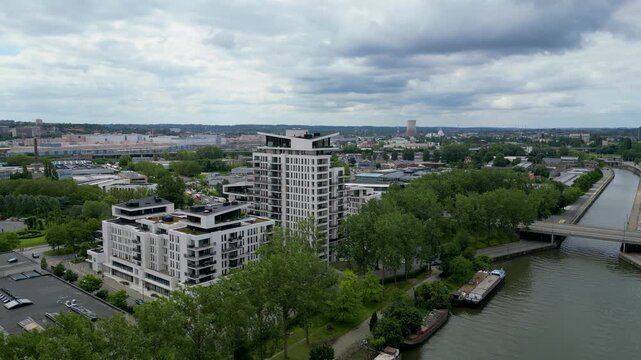 Aerial view of modern residential apartment buildings along the canal with barges and lush green trees under a cloudy sky in Anderlecht, Brussels, Belgium.