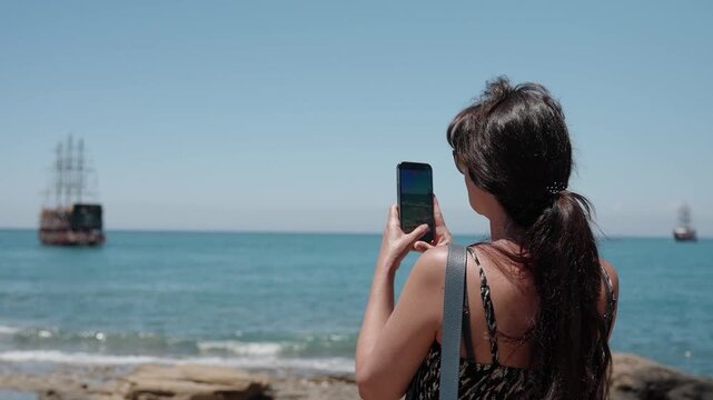 Woman tourist photographs the sea with smartphone on sunny beach, capturing sailing ship, travel memories, summer vacation, mobile photography and coastal leisure lifestyle.