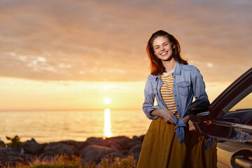 Fototapeta premium Cheerful woman outdoors, wearing denim jacket over striped top, sunset scene by the car, relaxed lifestyle moment, warm light, soft breeze, joyful expression, colors