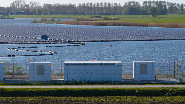Zwolle, Netherlands - 23 April 2026: Aerial view of a floating solar farm on a lake with white utility boxes in the foreground and green fields in the background.