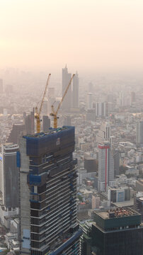 Bangkok, Thailand - 05 February 2026: Aerial view of a skyscraper under construction with two yellow cranes on top overlooking the dense urban skyline under a hazy morning sky.