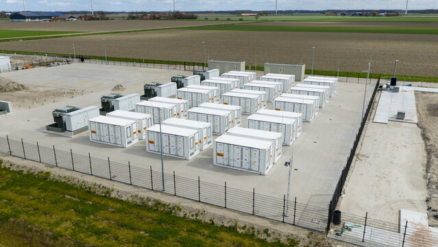 Dronten, Netherlands - 04 April 2026: Aerial view of a battery energy storage system facility with rows of white units surrounded by a fence and flat agricultural fields.
