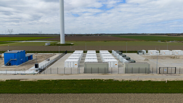 Dronten, Netherlands - 04 April 2026: Aerial view of a battery energy storage facility with rows of white containers and a wind turbine in a flat rural landscape.
