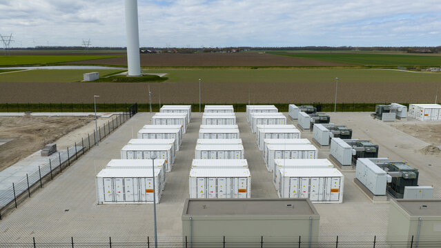 Dronten, Netherlands - 04 April 2026: Aerial view of a large-scale battery energy storage system with rows of white containers next to a wind turbine in a flat agricultural landscape.