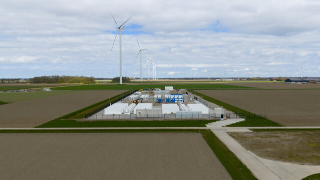 Dronten, Netherlands - 04 April 2026: Aerial view of a battery energy storage system facility surrounded by agricultural fields and wind turbines under a cloudy sky.