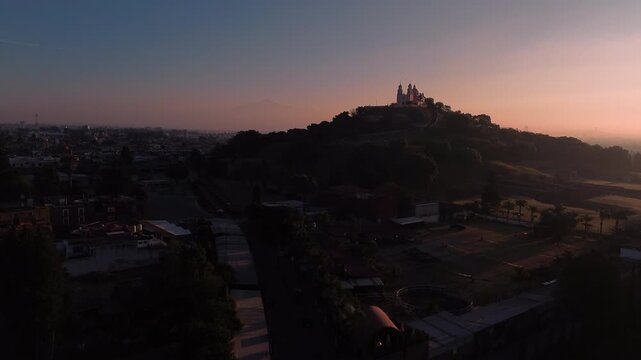 Aerial drone slowly approaches the Cholula pyramid and church at dawn, silhouetted against a magenta and blue sky with the Puebla city skyline and La Malinche volcano visible in the distance