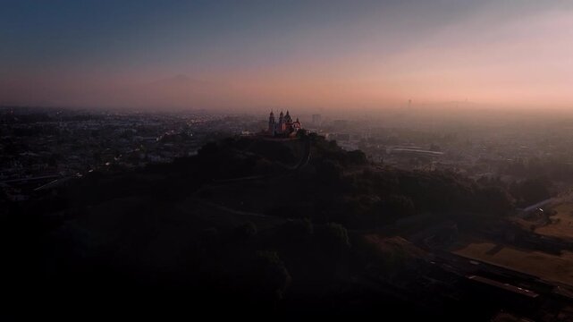 Aerial drone slowly approaches the Cholula pyramid and church at dawn, silhouetted against a magenta and blue sky with the Puebla city skyline and La Malinche volcano visible in the distance