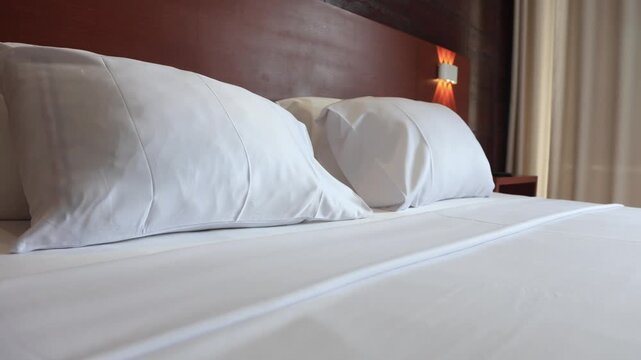 Panning across a double bed with white sheets, immaculately made in a minimalist and sober space. It looks like an elegant space, with background lights and a pleasant, even lighting.
