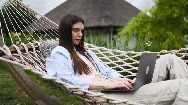 Young woman enjoying flexible remote work setup with laptop in hammock outdoors. Concept freedom, nature and productivity.
