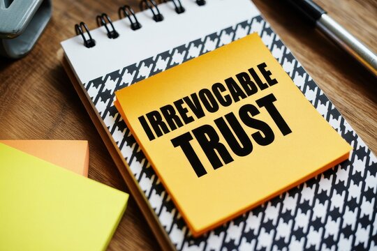 Yellow sticky note with Irrevocable Trust handwritten text placed on a spiral notebook on a wooden desk