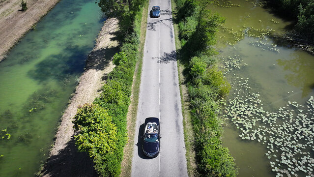 Succieu, France - 13 June 2026: Aerial view of two convertible cars driving along a narrow road between a canal and a pond with water lilies.