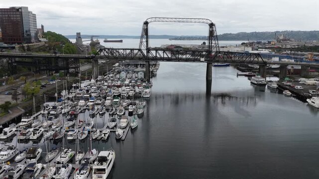 Packed marina with sailboats and yachts fills Thea Foss Waterway beneath historic Murray Morgan Bridge with cargo ship and Puget Sound beyond in Tacoma, Washington, USA. Aerial drone orbit wide shot