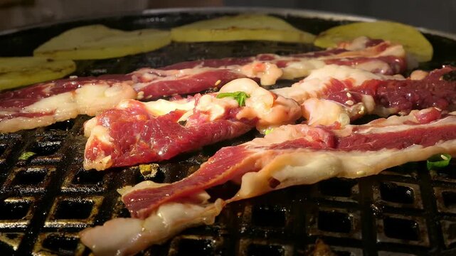 Juicy marinated beef slices sizzle on a Korean BBQ grill with golden potato slices, sesame seeds and fresh green scallions scattered atop