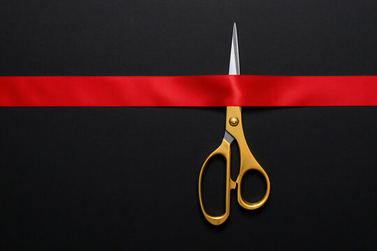 Grand opening. Top view of gold scissors cutting red silk ribbon against black background, copy space.