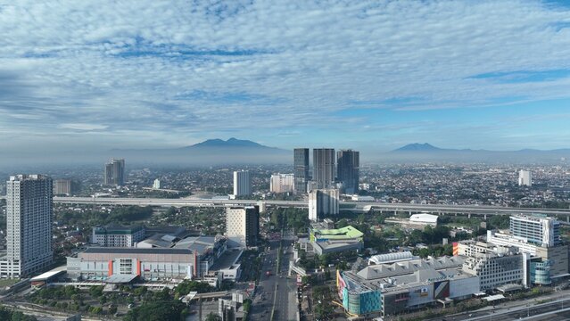Bekasi Barat, Indonesia - 12 April 2026: Aerial view of the urban skyline with high-rise buildings, highways, and mountains visible in the distance under a blue sky.