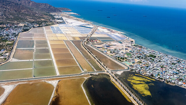 Drone aerial view of salt production fields near Ca Na, Ninh Thuan Province, Vietnam. The geometric grid of salt evaporation ponds reflects the sky in a mosaic of colours, with the coastal settlement 