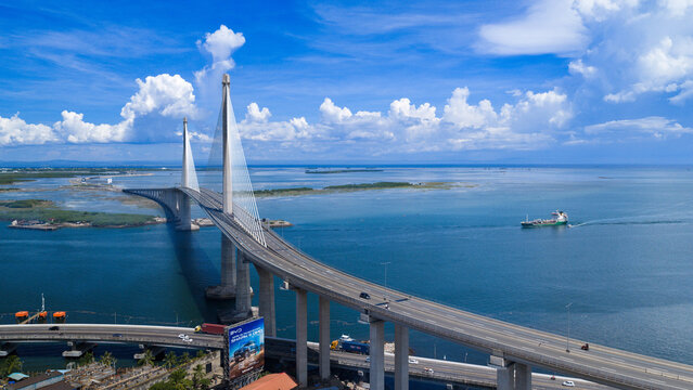 Cebu City, Philippines - 12 June 2026: Aerial view of the Cebu-Cordova Link Expressway bridge spanning the Mactan Channel with a cargo ship on the blue water under a cloudy sky.