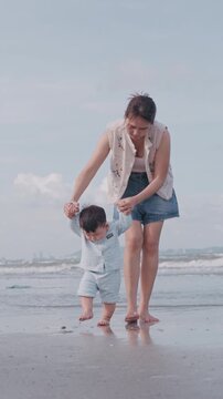 Young mother holding her baby's hands and helping him to take first steps on the sandy beach.