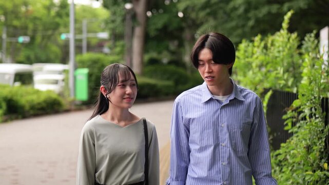 話しながら新緑の歩道を歩く若いカップルのトラッキングショット