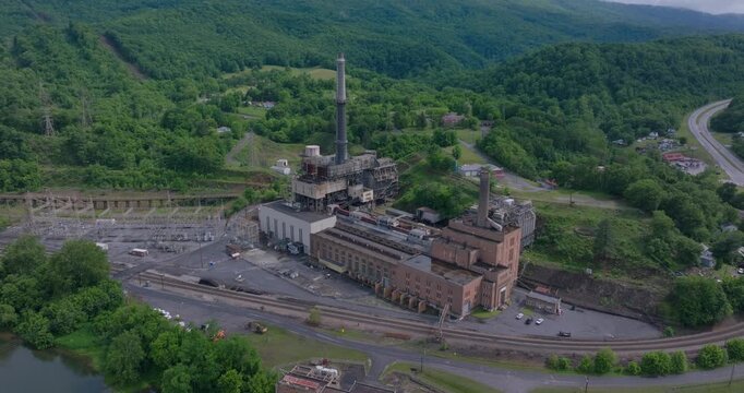 Industrial power plant complex surrounded by green Appalachian hills, drone medium tilt up pullback