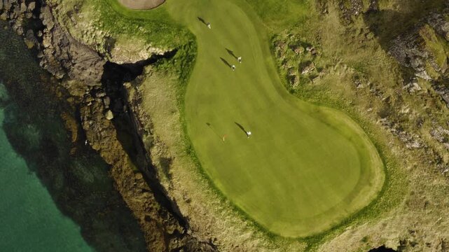 An aerial top-down view of people playing golf on a beautiful, green course situated on a rugged, rocky coastline next to the ocean.