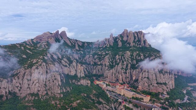 Santa Maria de Montserrat Abbey nestled below jagged conglomerate peaks with clouds drifting through the massif and the Catalan plain visible beyond, Catalonia, Spain