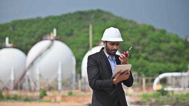 Engineers wearing safety gear, including hard hats examining survey a large blueprint tablet standing industrial facility gas or oil refinery engaged in a job requires high safety standards concept.	