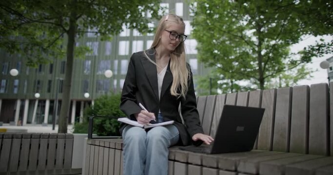 A young Caucasian female student wearing glasses in a modern urban environment among houses and trees sits on a bench in a park and works on a laptop writing notes in a notebook