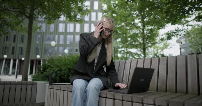 A young Caucasian female student wearing glasses in a modern urban environment among houses and trees sits on a bench in a park and works on a laptop and uses a mobile phone