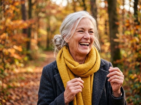 Laughing senior woman with hair bun walking on autumn forest path with falling leaves