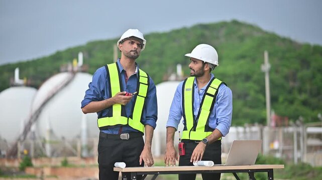 Engineers wearing safety gear, including hard hats examining survey a large blueprint tablet standing industrial facility gas or oil refinery engaged in a job requires high safety standards concept.	