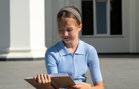 Smiling teen student girl reading book sitting on school building background on sunny day. Schoolgirl outdoors. Back to school concept