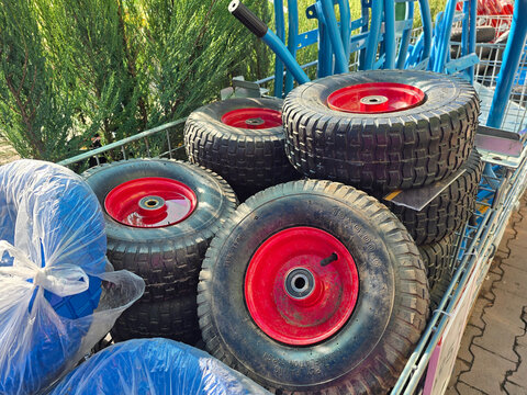 A pile of sturdy replacement wheelbarrow wheels with red hubs and black rubber tires is stacked in a metal cart outdoors at a garden center, ready for equipment repair, landscaping, or utility use
