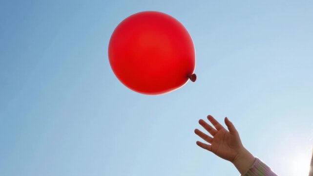 Red rubber ball floating in air against clear blue sky with sun flare and human hand reaching up to catch it during daytime outdoor activity