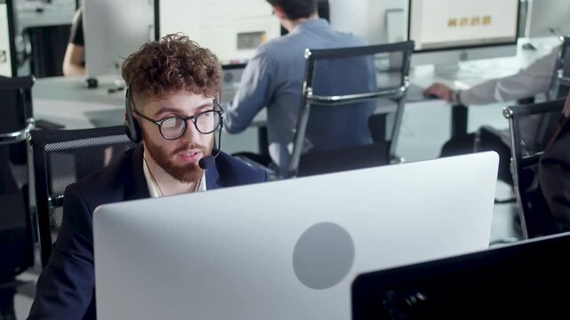 Young male customer support agent talking through headset while working at computer in a modern open plan office. Professional call center employee provides online assistance and communicates with