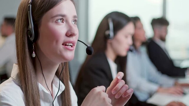 Young female customer support agent wearing headset and smiling while speaking with client in a modern call center office. Professional support operator gestures with hands during conversation