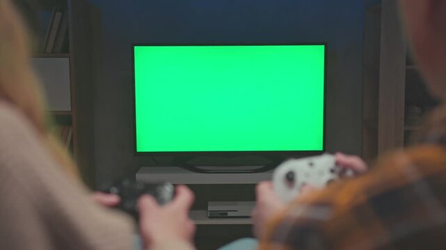 Chroma Key Television Screen Mockup with Blurred Gamers Holding Controllers. Unrecognizable Young Couple Plays Virtual Video Game Template on Electronic Console. Creative Media Production Concept 4k