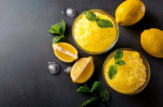 Two bowls of lemon granita sit on a dark table. Fresh mint leaves are placed on top. Whole lemons and lemon halves surround the bowls. Ice cubes are scattered nearby.
