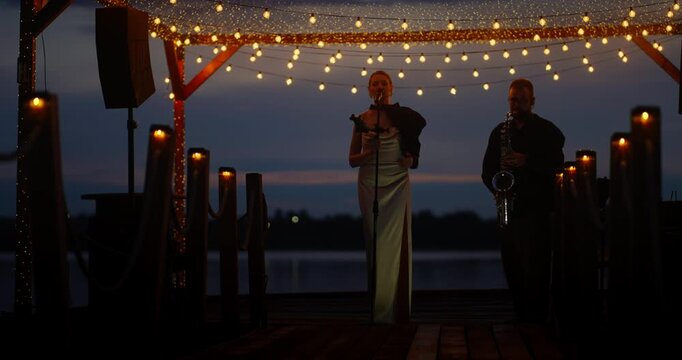 Female singer and saxophonist performing on outdoor wooden stage at dusk