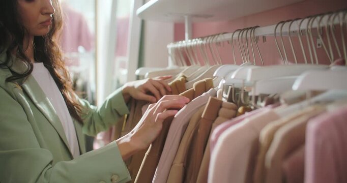 Stylish young woman browsing clothes on a rack in a modern fashion boutique. Female shopper in a pastel blazer touches garments and selects outfit options in a soft retail interior with curated