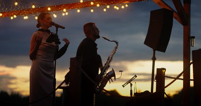 Female singer and barefoot saxophonist on wooden outdoor stage at dusk