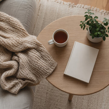 Cozy cocooning lifestyle scene with warm knitted blanket, cup of tea, book and green plant on wooden table in living room