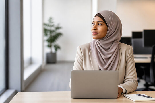 A Muslim woman wearing a hijab sits at a desk using a laptop in a bright, modern office setting.