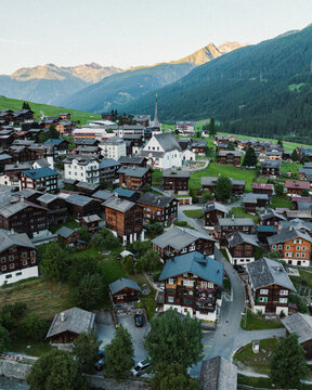 Goms, Switzerland - 09 August 2024: Aerial view of the village of Munster featuring traditional wooden chalets and the white church of St. Mary surrounded by green meadows and the Swiss Alps.