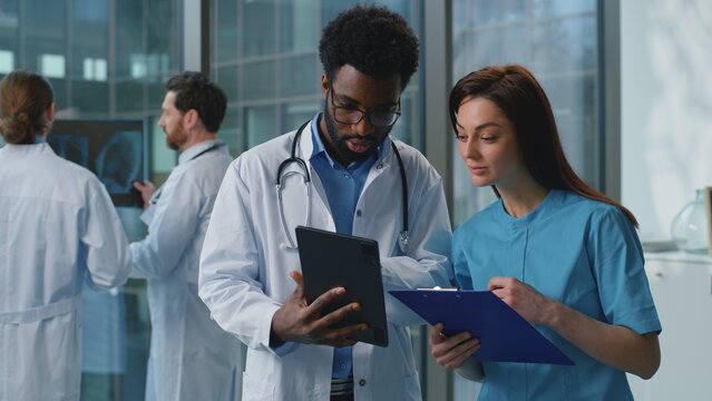 Two diversity doctors man with woman discussing medical examination using digital tablet together in hospital. Caucasian female therapist doctor talking with African American male surgeon at clinic