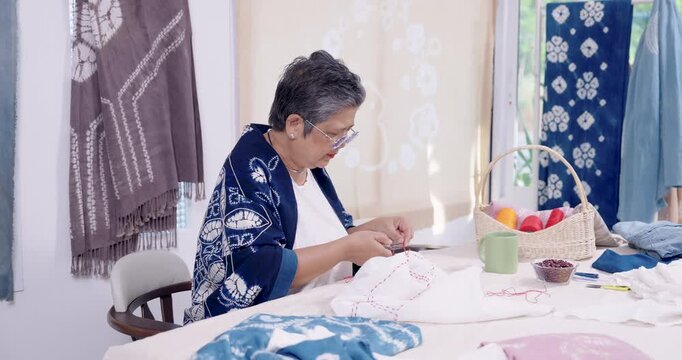 Retired Asian woman entrepreneur bends close, marking and stitching resist pattern on cloth, preparing handmade indigo dye textile for customer order, slow craft workshop at home studio, custom.